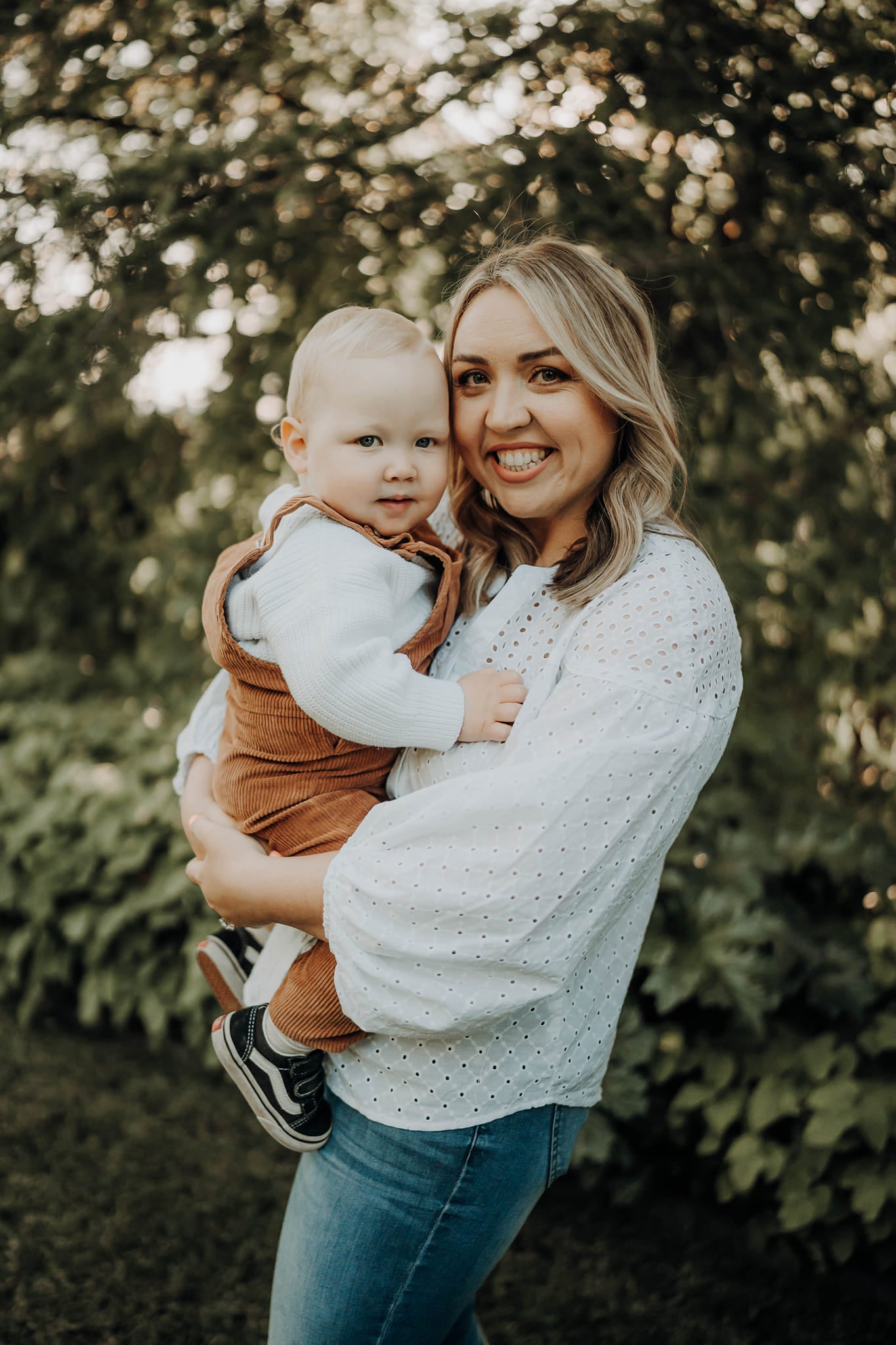 An image of Motherhood Milestones founder, Emily Mitchell, holding her son. Both are looking and smiling at the camera. Emily is wearing a white long sleeve shirt and jeans and her son is in brown overalls and a white jumper. They are outside at sunset in front of trees.