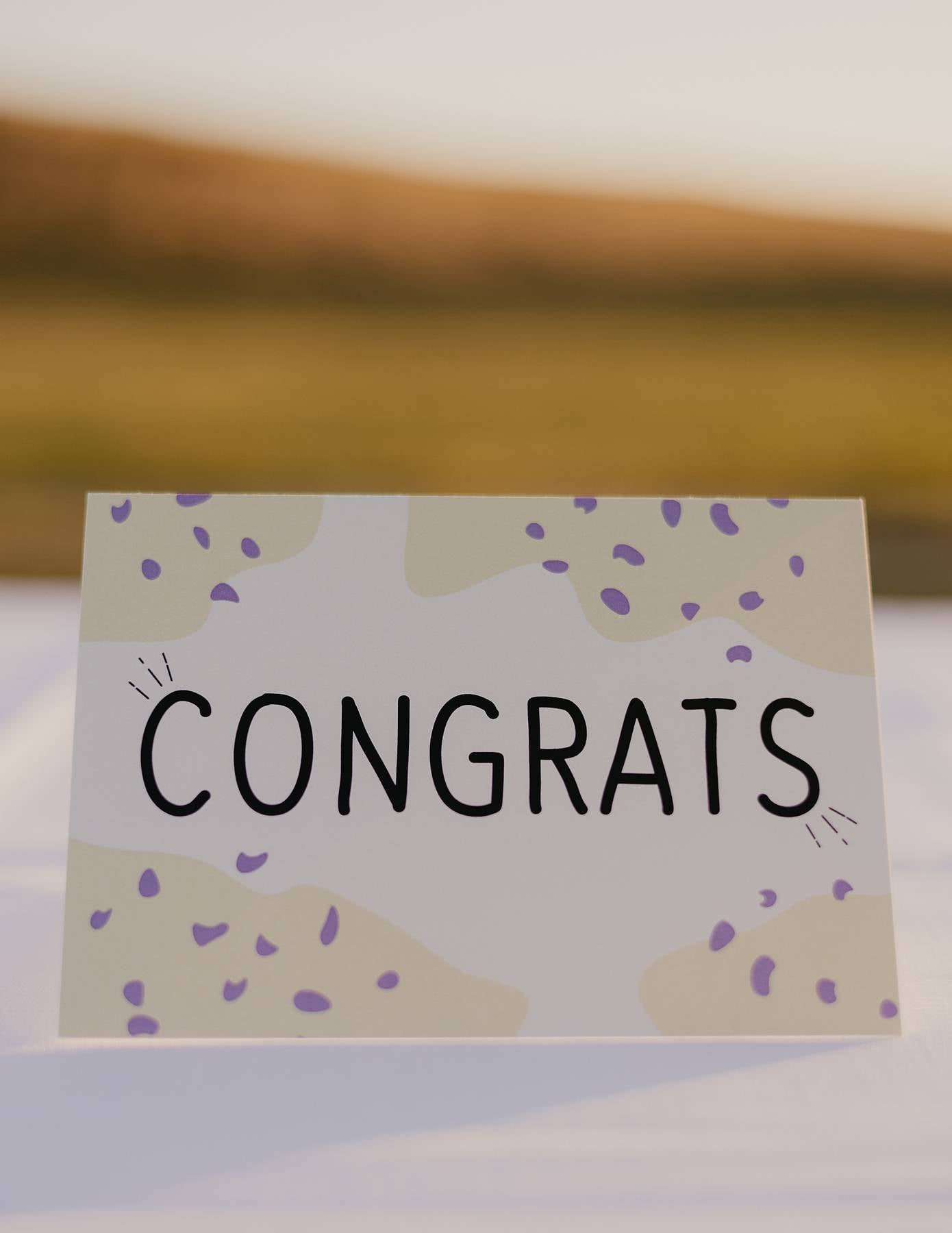 The front of a Motherhood Milestones card that reads Congrats and has a yellow background with darker yellow splotches in the corner with purple dots. Congratulations.