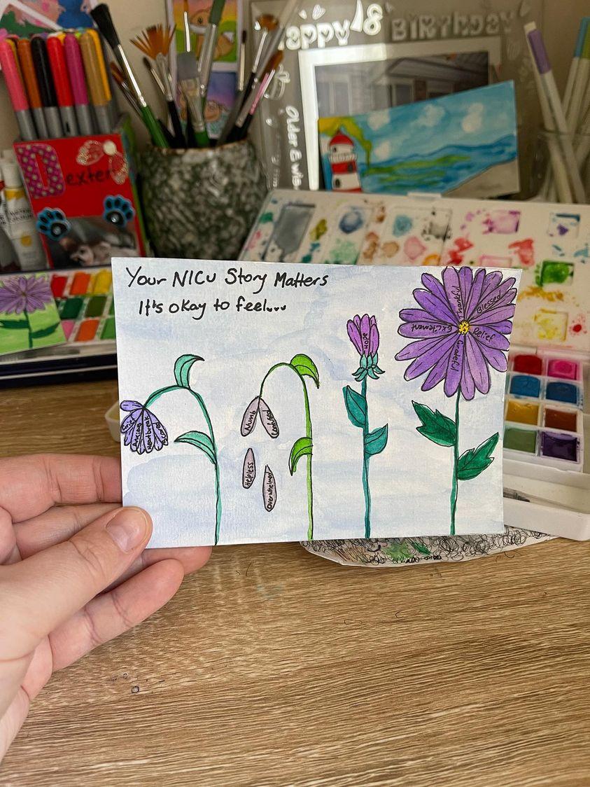 A hand holding a water colour painting of the NICU mum card, with four flowers next to each other at different stages of growth with petals containing the words of different emotions mothers feel through the NICU experience. The painting is being held by a card and the background is a table full of art supplies.