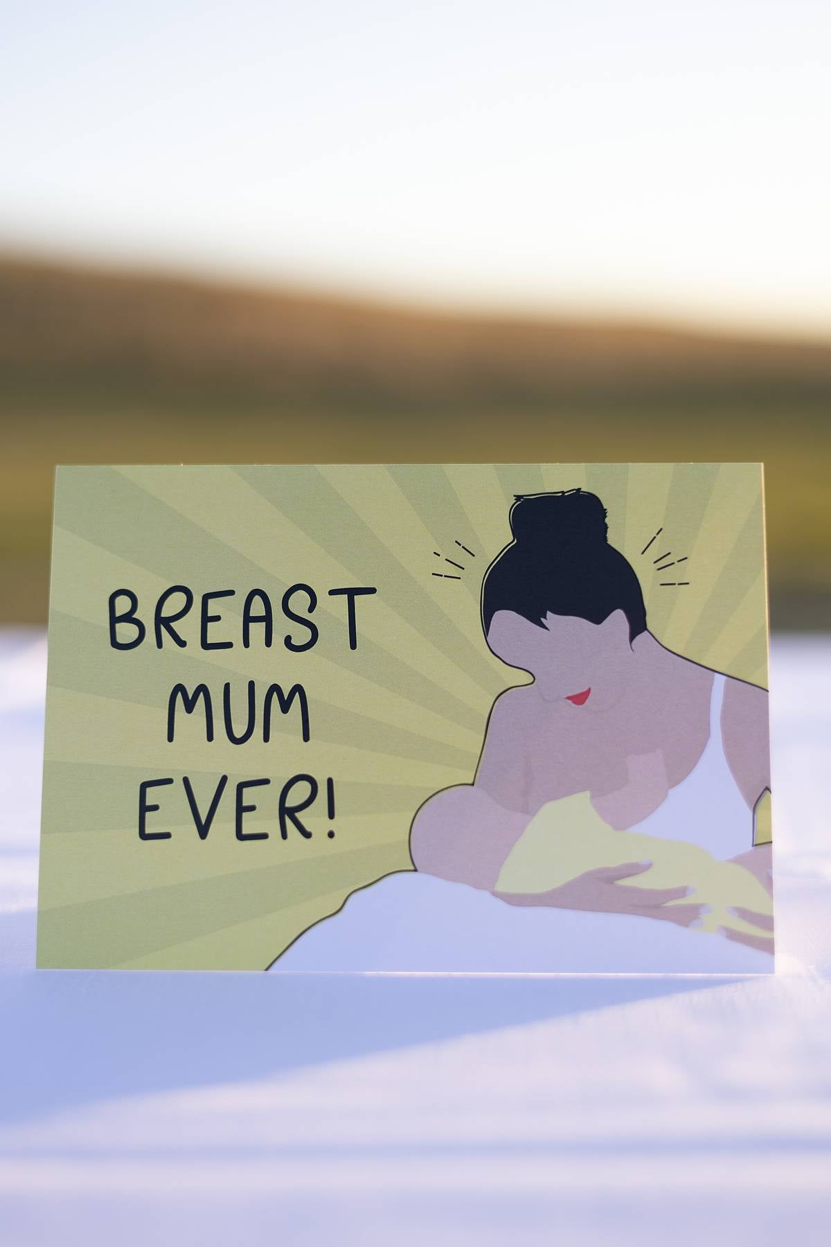 Front of a Motherhood Milestones card which reads Breast Mum Ever, celebrating a mothers breastfeeding or weaning journey. There is a picture of a mum breastfeeding next to the words and a background of yellow rays like a sun.