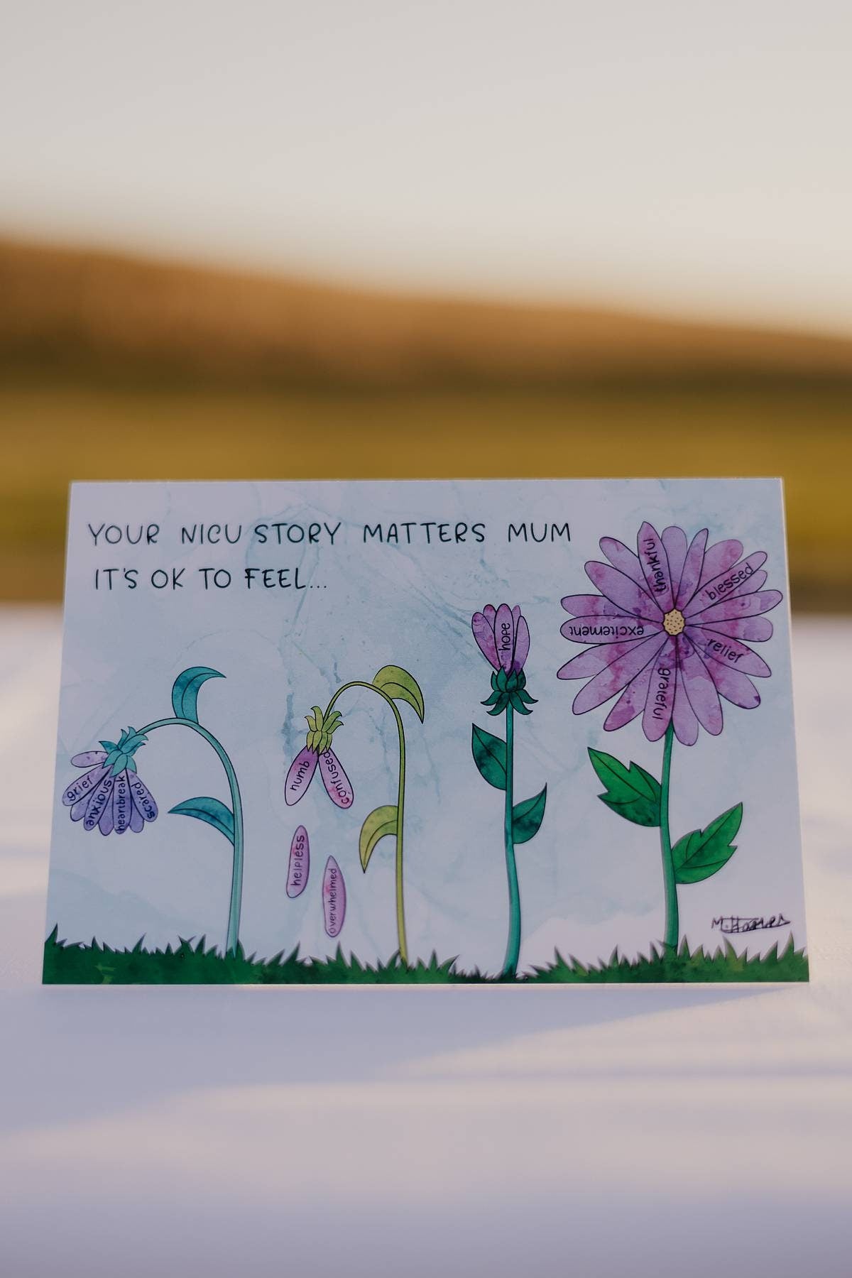 A hand holding a water colour painting of the NICU mum card, with four flowers next to each other at different stages of growth with petals containing the words of different emotions mothers feel through the NICU experience. The painting is being held by a card and the background is a table full of art supplies.