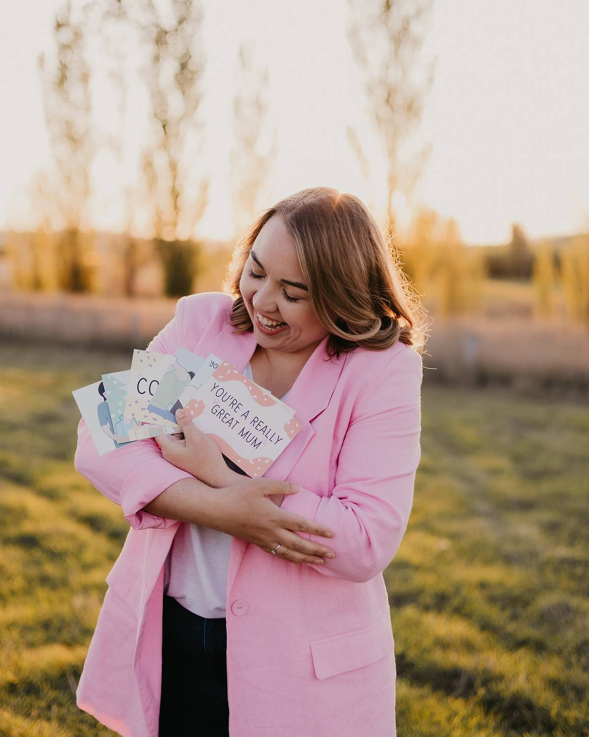 Founder of Motherhood Milestones, Emily Mitchell, cradling a selection of her Motherhood Milestones cards. She is outside at sunset, wearing jeans, a white shirt and a pink blazer. 