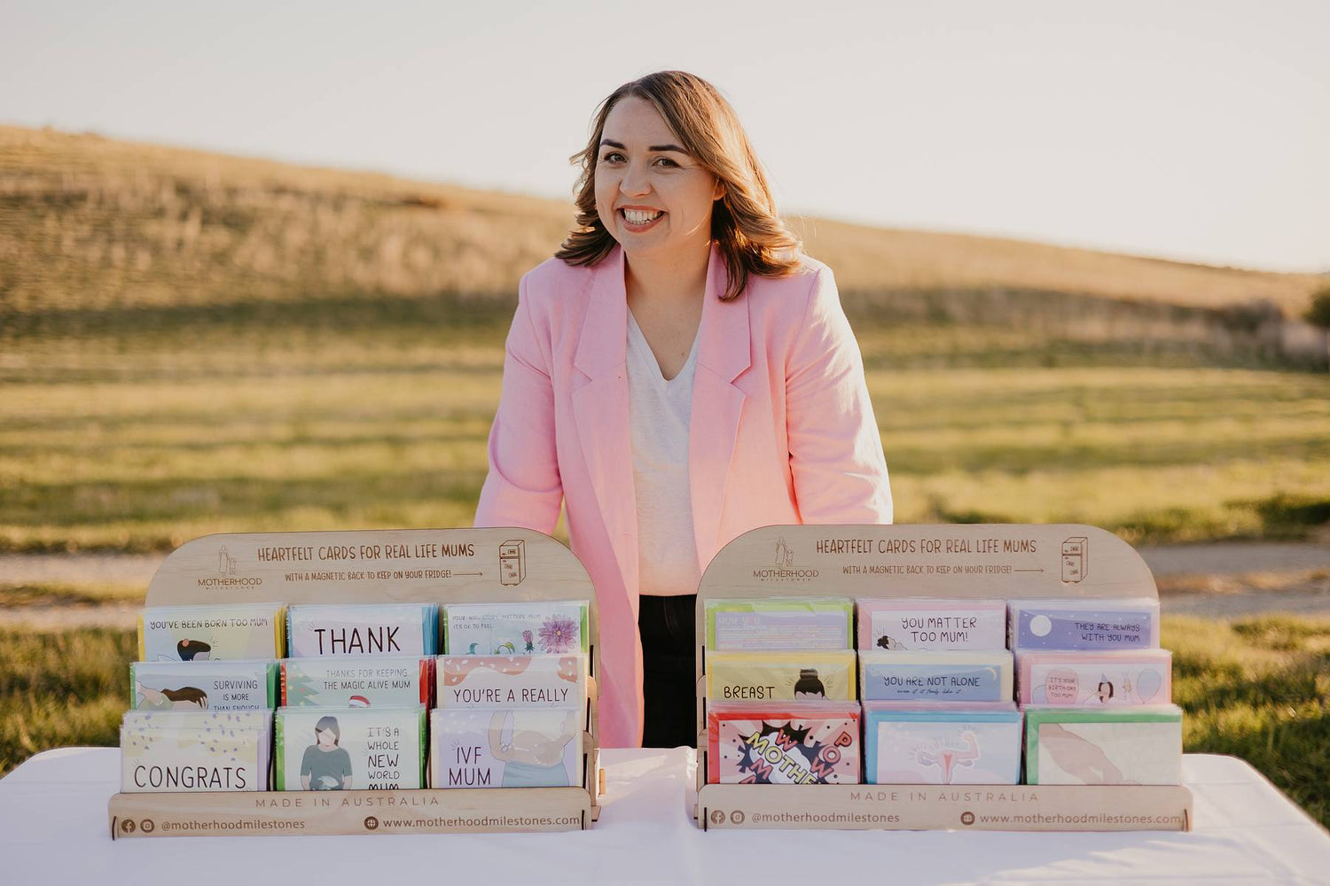 Founder of Motherhood Milestones, Emily Mitchell, standing up behind a table with a white tablecloth. On the table are two wooden Motherhood Milestones card stands filled with each design from the Motherhood Milestone collection, all celebrating or commiserating moments in Motherhood. 
