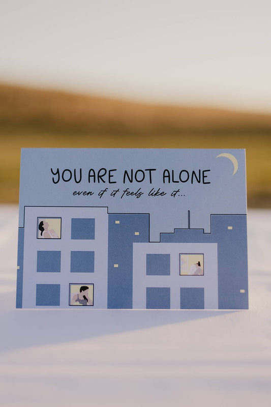 Font of motherhood milestones card that reads you are not alone even if it feels like it. There is an image of buildings with come window lights on and mothers and babies awake throughout the night. 