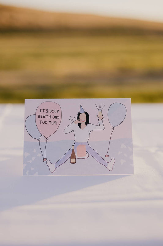 The front of a Motherhood Milestones card that shows a mum on the floor with a champagne in her hand, balloons all around her and a cake in front of her, reminiscent of a cake smash for a toddler but instead celebrating the mother and all she has been through and achieved in that year of parenting and motherhood. 