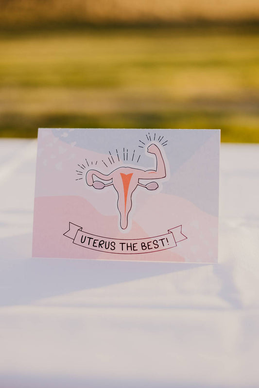 The front of a Motherhood Milestones card that reads uterus the best with a picture of a uterus flexing its muscles (tubes). The background is dark pink, light pink and blue. 
