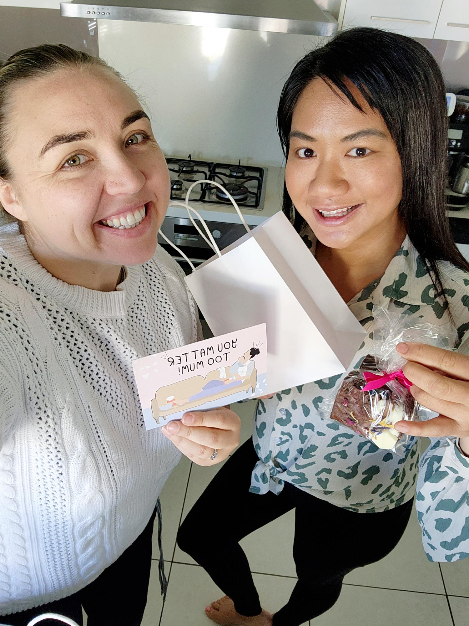 A photo of two mothers smiling and holding up a Motherhood Milestones card, a paper gift bag and brownies making up a Mumma Care Package to celebrate mothers.