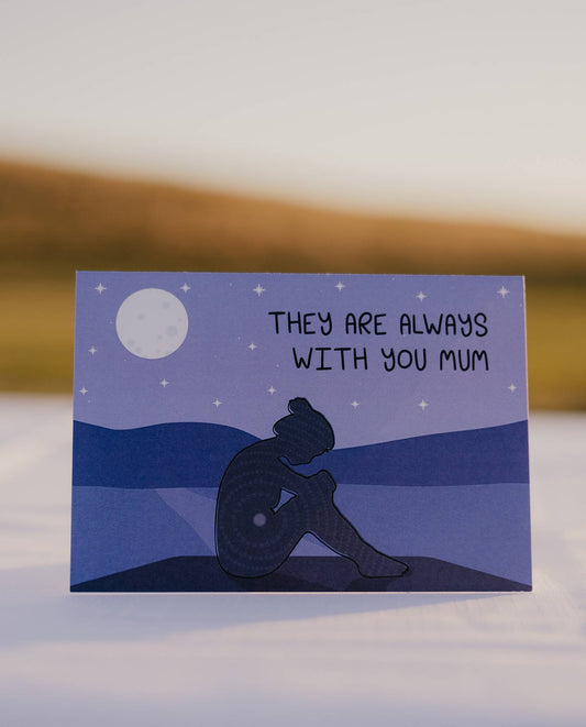 Front of a motherhood milestones card that reads they are always with you mum. There is a mother sitting on her bottom holding her knees up with her hands with her head bent forward looking down. Behind her is water, mountains and the night sky with a moon.