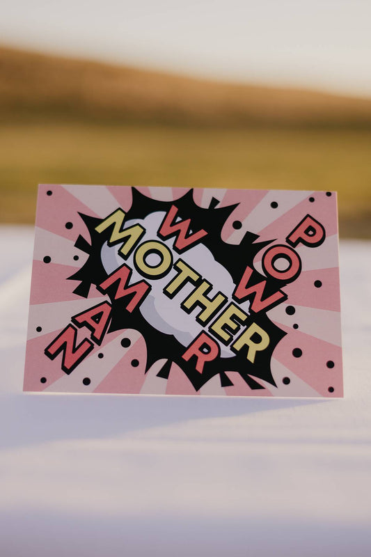 The front of a Motherhood Milestones card that reads Mother with the words Power and woman coming off it. There is a super hero like background with a black section with a cloud and lines of dark pink and light pink to represent a punch.