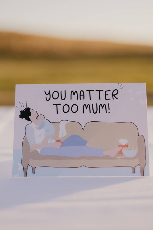 The front of a Motherhood Milestones card that reads you matter too mum. Pictured is a mum on a lounge holding a newborn baby with her head back, feet up and coffee cup in hand.