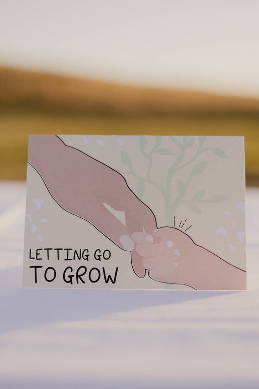 The front of a Motherhood Milestones card that has a picture of a mothers hand holding a toddlers hand and the words letting go to grow to represent times of transition and separation in the motherhood journey.