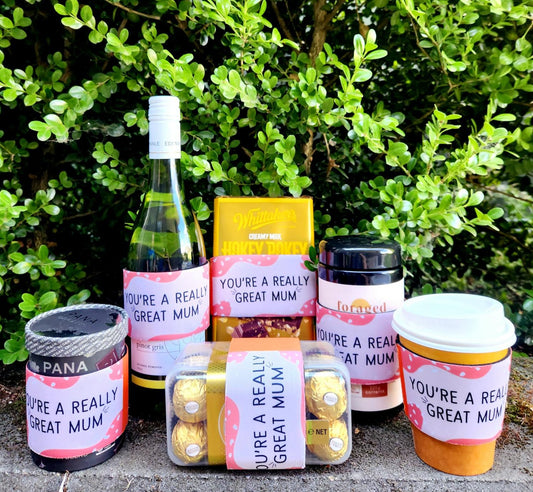 Six products including, wine, chocolate, chocolate powder, supplements, and a coffee cup are sitting on a grey fence with a green leafy background. All products have a Motherhood Milestones neoprene sleeve on them that says you're a really great mum, in an orange/pink tone. 