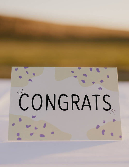 The front of a Motherhood Milestones card that reads Congrats and has a yellow background with darker yellow splotches in the corner with purple dots. Congratulations. 