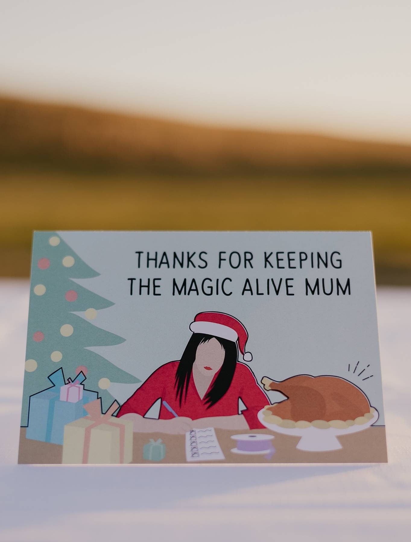 The front of a Motherhood Milestones card that pictures a mother in a santa hat at a table checking a list. Next to her are presents and a turkey and to her side is a christmas tree. The text reads thanks for keeping the magic alive mum. This card is an ode to all mothers do for the holidays. 