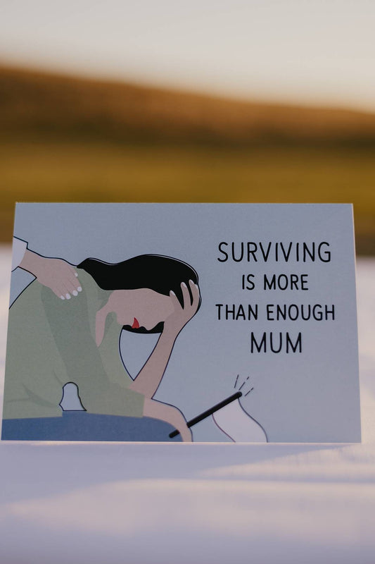 The front of a Motherhood Milestones card picturing a mum with her head in her hands, holding a white flag. There is a hand on her back and writing on the card that says surviving is more than enough mum. The background is blue. This card acknowledges that motherhood is hard and sometimes surviving the day is more than enough to maintain your mental health.