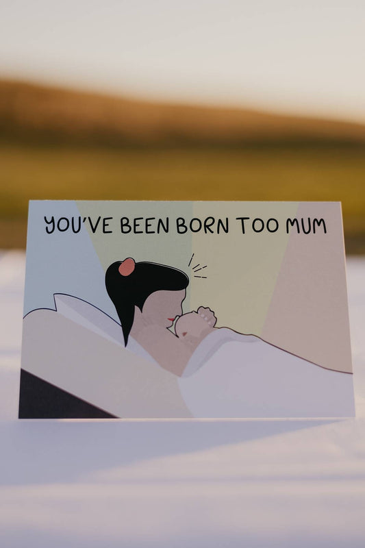 The front of a Motherhood Milestones card which reads You've been born too mum, celebrating a womans journey into motherhood (matrescence). The mother is on a bed cradling her newborn and their are four colours filling the background, representative of each season.