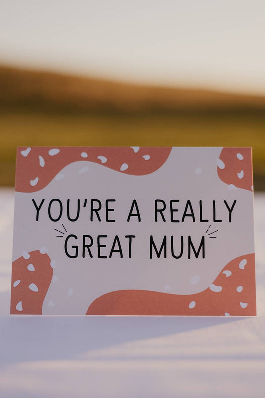 The front of a Motherhood Milestones Card that reads you're a really great mum in black text. The card had a light pink background with orange splotches in corners with white dots over the top.  